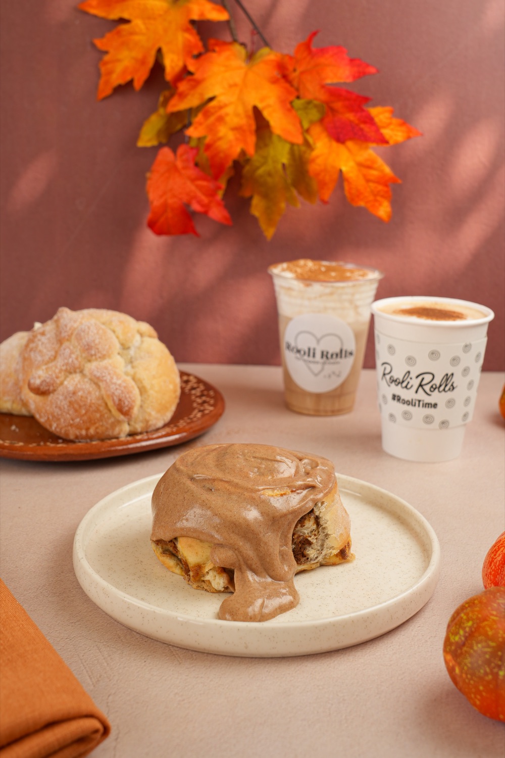 Pan de muerto y Rol Pumpkin Spice servidos junto a bebidas Rooli Rolls, con hojas de otoño y calabazas